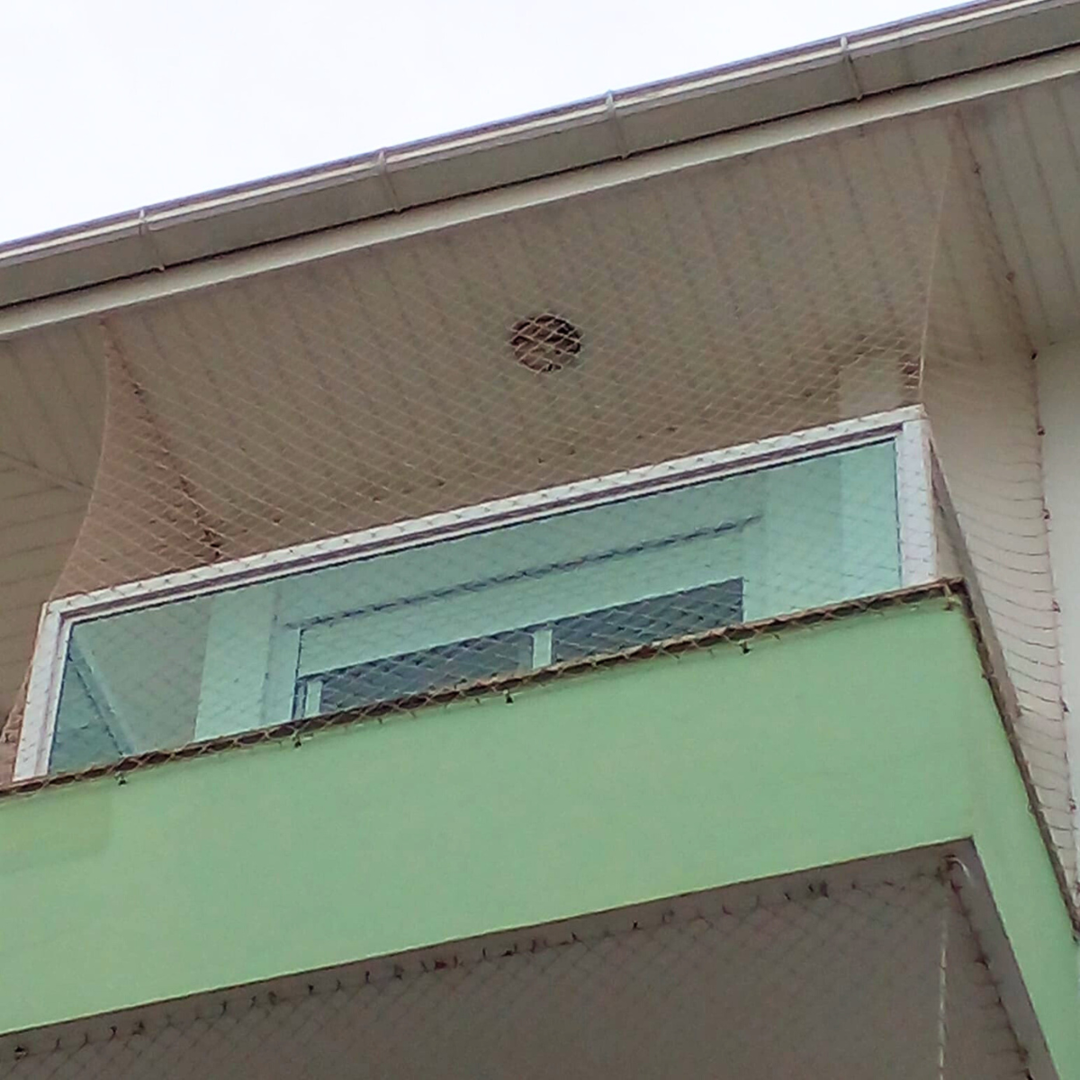 Vista inferior de sacada com guarda-corpo de vidro verde, totalmente fechada com rede de proteção até o beiral do telhado.