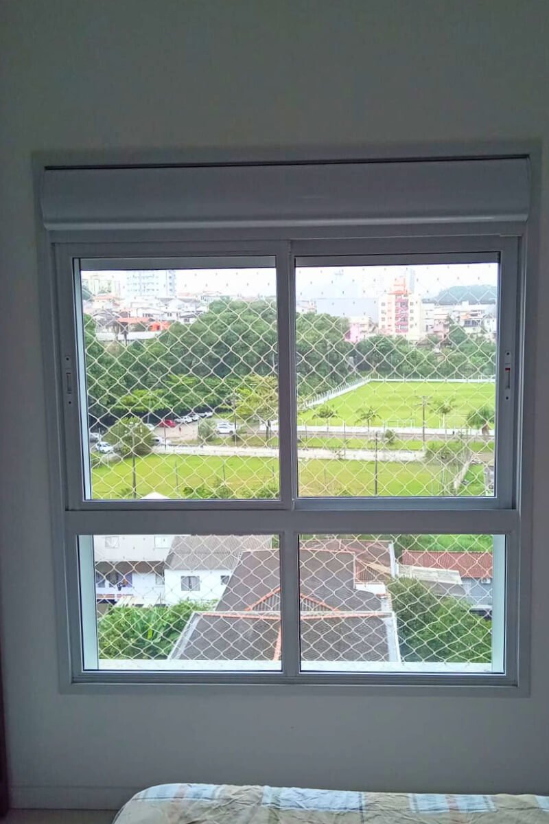 Janela de alumínio de correr com rede de proteção, permitindo a vista segura para um campo de futebol gramado ao fundo.