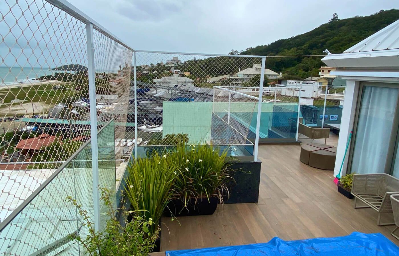 A segurança é feita com estrutura metálica branca e rede fixada sobre o guarda-corpo de vidro, com vista para o mar ao fundo.