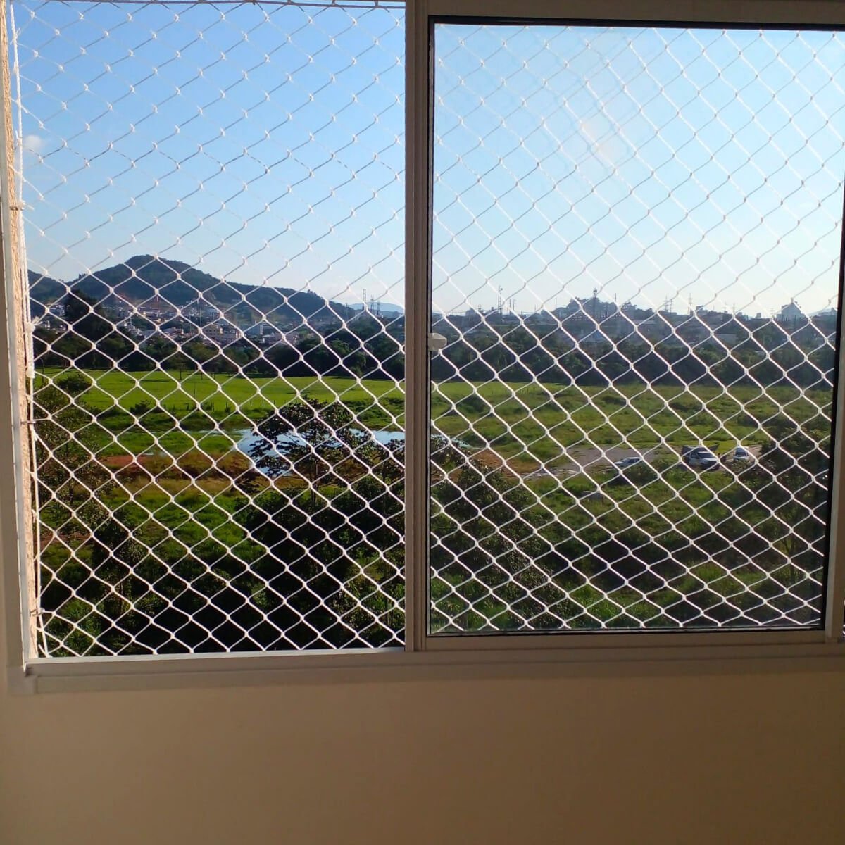 Janela residencial com rede de segurança instalada, oferecendo vista ampla para área verde e montanhas.
