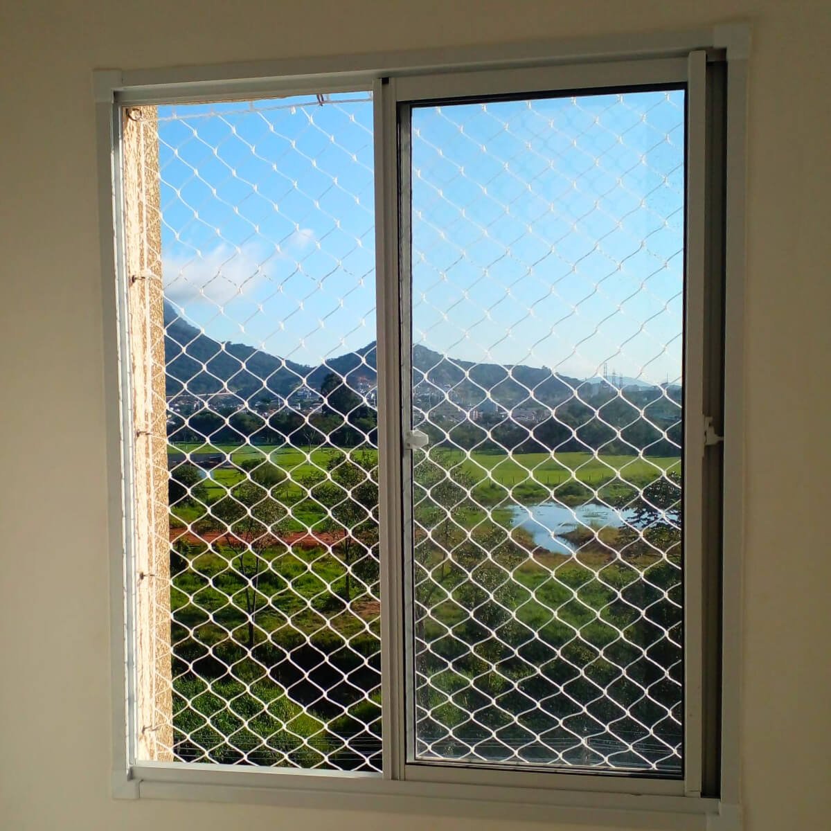 Janela de quarto com rede de proteção e vista para paisagem natural e céu azul.