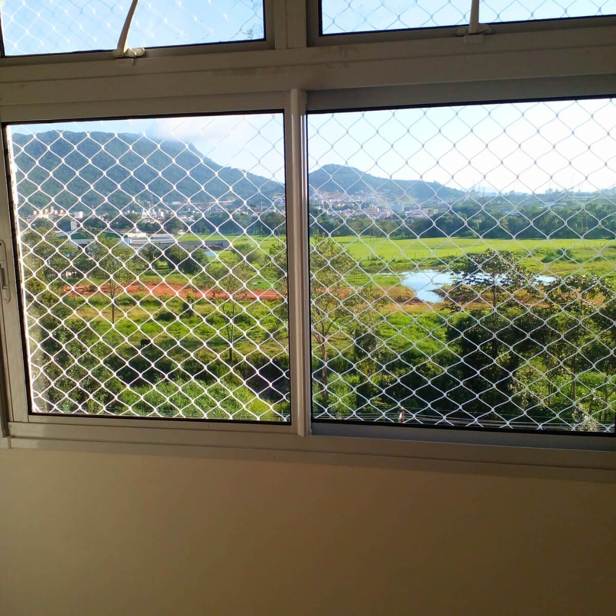 Janela de correr com rede de proteção instalada, com vista para vegetação densa e morros ao fundo.