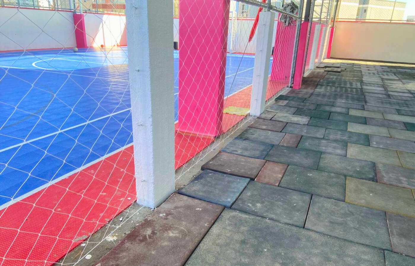 Quadra poliesportiva com piso azul e vermelho cercada por rede de proteção branca sustentada por pilares rosa.