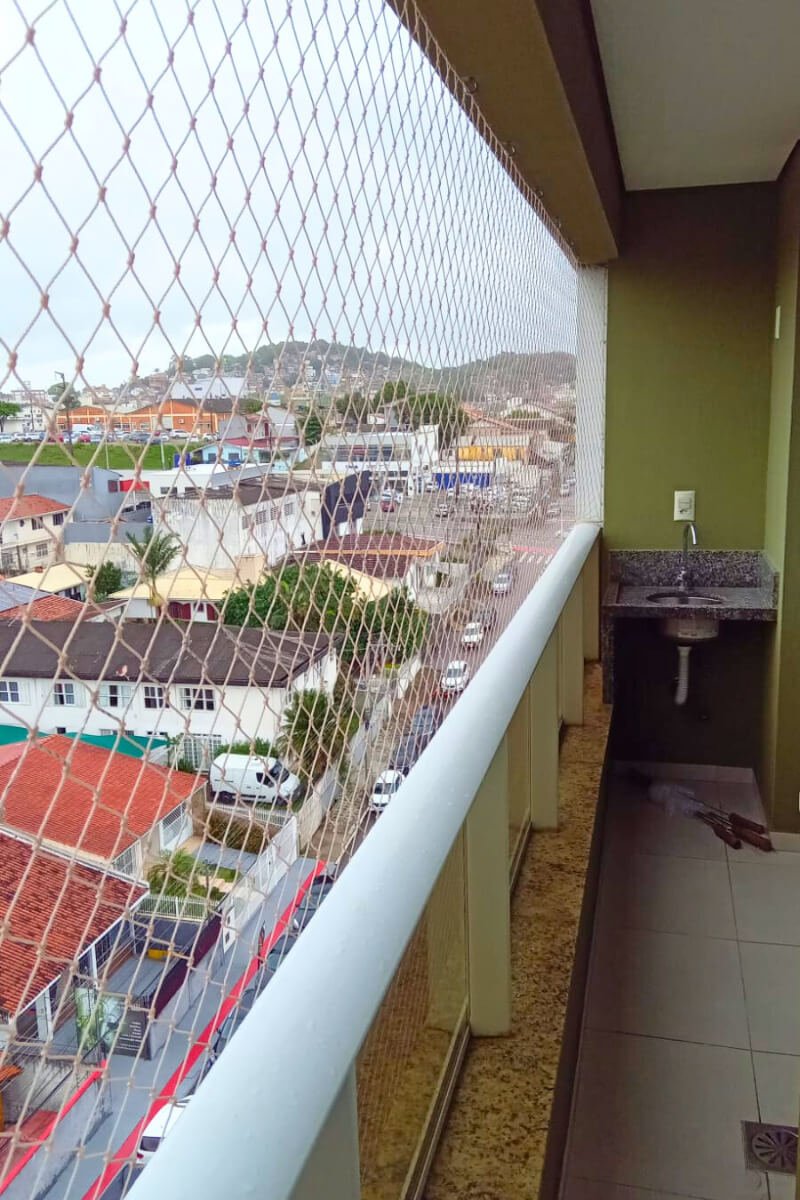Vista superior de sacada com guarda-corpo de vidro e fechamento total em rede de proteção, com vista para o bairro.